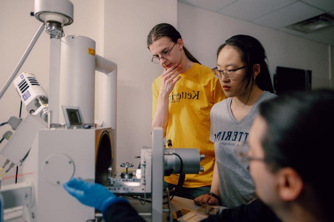 A Kettering professor is seated at an electron microscope instructing two students standing nearby